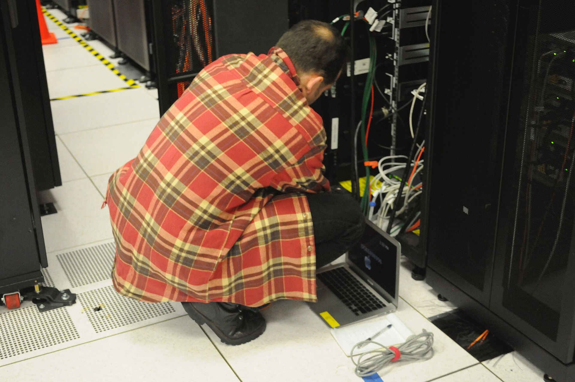 A technician kneels on a raised data-centre floor, working on a server rack with a laptop.